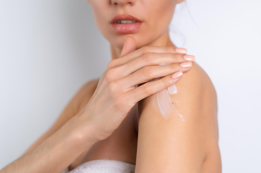 A beauty portrait of a topless woman with perfect skin, holding an eczema-safe bottle of lotion and applying it to her shoulder.